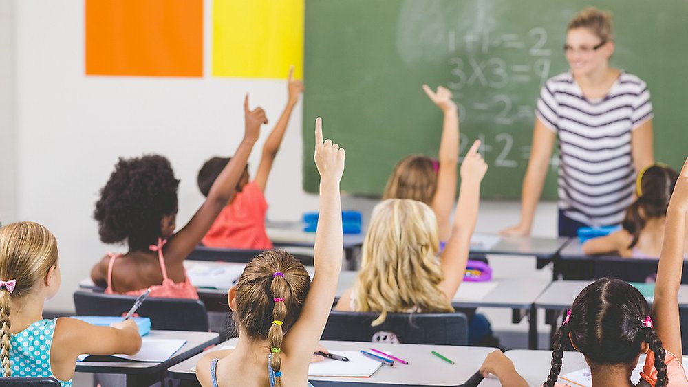 Skolelever i klassrum räcker upp handen